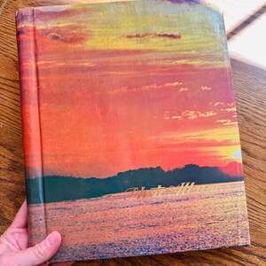 May include: A vintage hardback book with a sunset scene on the cover. The cover features a colourful sky with orange, yellow, and pink hues, and a dark silhouette of trees and water in the foreground. The book's title is visible in gold lettering at the bottom of the cover.