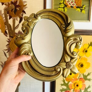 May include: A gold-colored, oval-shaped mirror with a decorative floral frame. The frame features ornate details and flower accents. The mirror is held up against a backdrop of framed floral embroidery.