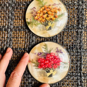 May include: Two circular coasters featuring pressed dried flowers arranged in a bouquet inside a black and gold decorative basket. The top coaster displays orange flowers, while the bottom coaster features vibrant red flowers.