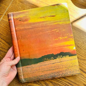 May include: A vintage photo album with a sunset scene on the cover. The cover is orange, yellow, and red with a blue lake and green trees in the foreground. The words "Photo Album" are printed in gold on the cover.
