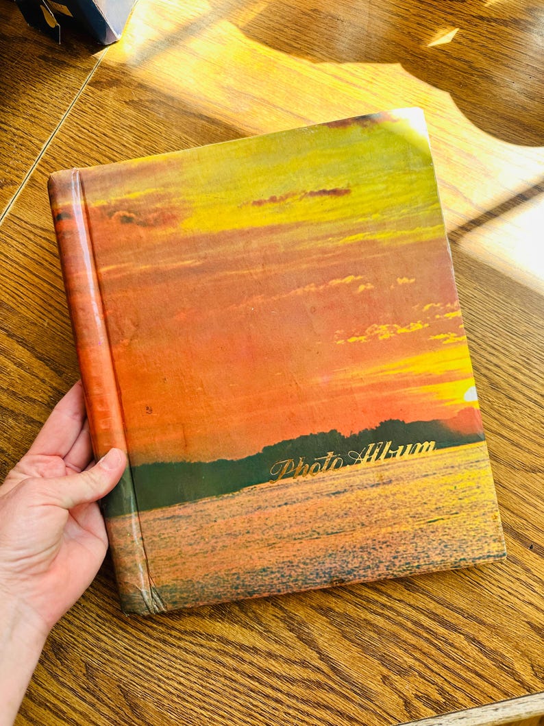 May include: A vintage photo album with a sunset scene on the cover. The cover is orange, yellow, and red with a blue lake and green trees in the foreground. The words "Photo Album" are printed in gold on the cover.