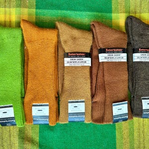 Peut inclure: Cinq paires de chaussettes de couleur verte, orange, beige, marron et grise, disposées sur une surface à carreaux verts et jaunes. Chaque paire porte une étiquette "Interwoven" et "Crew-Sader".