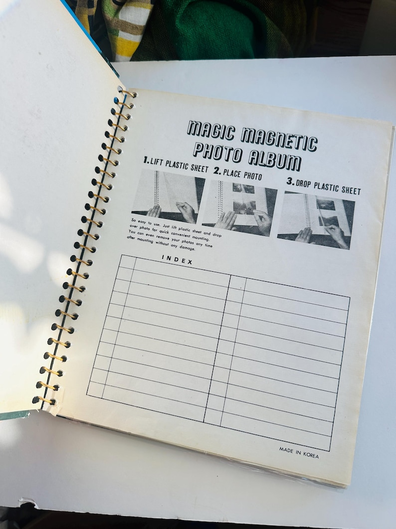 May include: A black and white photo album with a gold spiral binding. The cover has the text "Magic Magnetic Photo Album" and instructions for using the album: "1. Lift Plastic Sheet 2. Place Photo 3. Drop Plastic Sheet". The album has an index page with lines for writing in.
