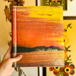 May include: A vintage photo album with a sunset scene on the cover. The cover is orange, yellow, and red with a dark silhouette of trees and water. The words "Photo Album" are printed in gold lettering on the cover.