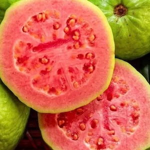May include: Close-up of four guava fruits, two whole and two cut in half, revealing the pink flesh and black seeds. The guavas are arranged on a woven surface with green leaves in the background.