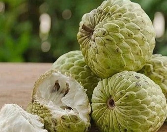 Könnte beinhalten: Nahaufnahme von mehreren grünen Custardäpfeln, einige mit entfernter Schale, die das weiße Fruchtfleisch im Inneren freilegen. Die Früchte sind auf einer Holzoberfläche angeordnet.