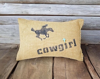 May include: A burlap pillow with a black silhouette of a horse and the word "COWGIRL" printed on it. A small turquoise cross is also visible on the pillow.