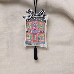 May include: A red house-shaped fabric sachet with a black and white gingham bow, a black tassel, and the text "This hut is my home, you're welcome here."
