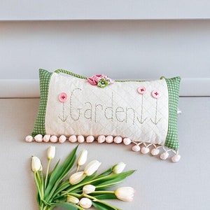 May include: A rectangular pillow with the word "Garden" embroidered on it, accented with pink buttons and a floral embellishment. The pillow has a green and white gingham border and is decorated with pink pom-poms. A bouquet of white tulips is in the foreground.