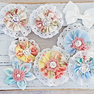 May include: Assortment of handmade fabric flowers with lace trim. The flowers are in various colors and patterns, including pink, blue, yellow, and green. Some flowers have a button or rhinestone center.