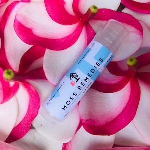 Könnte beinhalten: Ein weißer Lippenbalsam-Stift mit blauem und rotem Text, der "Naturally Perfect Moss Remedies Lip Balm For Sensitive Skin 4.4ml" lautet. Der Stift liegt auf einem Bett aus rosa und weißen Blütenblättern.