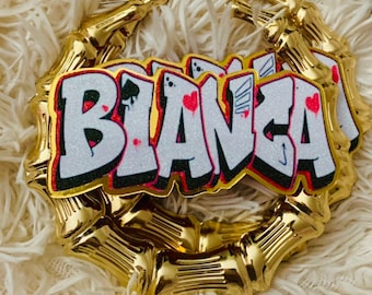 May include: Gold bamboo hoop earrings with a white and pink glitter 'BIANCA' nameplate. The nameplate has a graffiti style font with red hearts and playing card symbols.