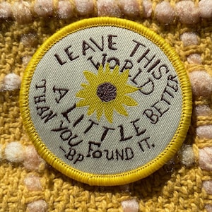 Może przedstawiać: Okrągła naszywka z żółtą obwódką i napisem "LEAVE THIS WORLD A LITTLE BETTER THAN YOU FOUND IT" otaczającym żółtą słonecznik. Naszywka znajduje się na teksturowanej żółtej tkaninie.