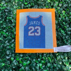 May include: A square needlepoint canvas featuring a blue basketball jersey design with the name JAMES and the number 23 printed in white. The canvas has a bright orange border and a small white fabric tag labeled Sunset Stitching.