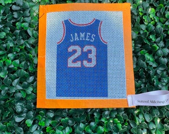 May include: A square needlepoint canvas featuring a blue basketball jersey design with the name JAMES and the number 23 printed in white. The canvas has a bright orange border and a small white fabric tag labeled Sunset Stitching.