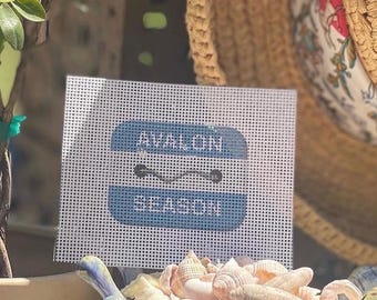 May include: A white needlepoint canvas featuring a rectangular blue graphic with the text AVALON and SEASON in white, separated by a thin black horizontal line with two small circles at each end, positioned above a collection of various seashells.