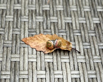 May include: A pair of round cufflinks featuring a woven fabric top with a multicolored pattern of orange, green, and tan threads, set in a polished silver-tone metal frame, resting on a single dried brown autumn leaf.