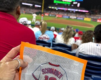 May include: A square needlepoint canvas featuring a white baseball jersey design with red trim, the name SCHWARBER, and the number 12 in red lettering. The canvas has a bright orange fabric border and is held up at a professional baseball stadium.