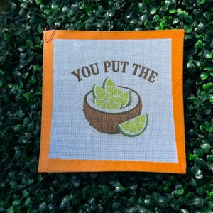 May include: A square needlepoint canvas featuring a brown coconut shell filled with light green lime wedges. The text YOU PUT THE is printed in brown above the coconut. The canvas has a bright orange border on all four sides of the white mesh.