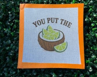 May include: A square needlepoint canvas featuring a brown coconut shell filled with light green lime wedges. The text YOU PUT THE is printed in brown above the coconut. The canvas has a bright orange border on all four sides of the white mesh.
