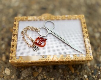 May include: A pair of silver-toned metal embroidery scissors with a gold-toned chain attached to the finger loop. The chain features a small, brown, pretzel-shaped charm with white dots and a silver-toned metal charm with the letters LB.