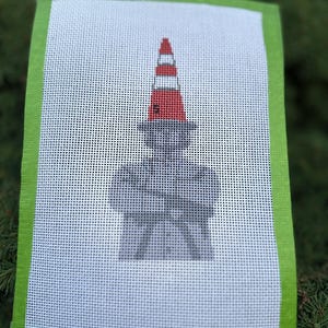 May include: A rectangular needlepoint canvas featuring a gray silhouette of a person with arms crossed, wearing a hat topped with a red and white traffic cone. The canvas has a bright green border and a small white label reading Sunset Stitching.