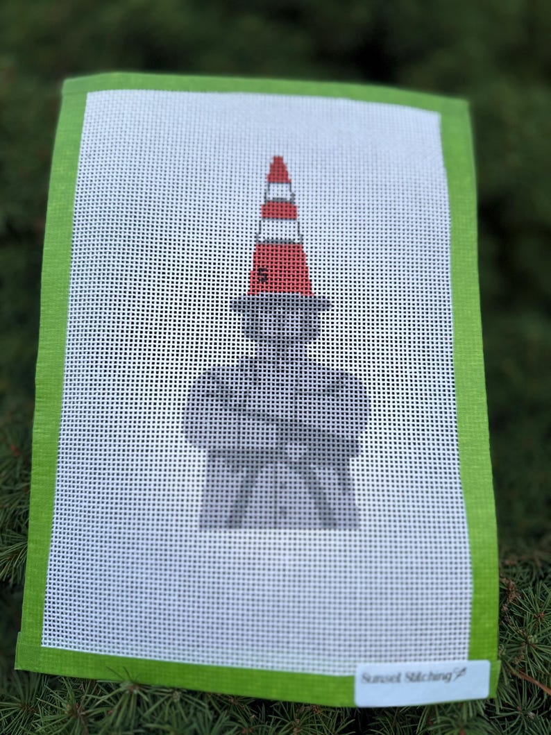 May include: A rectangular needlepoint canvas featuring a gray silhouette of a person with arms crossed, wearing a hat topped with a red and white traffic cone. The canvas has a bright green border and a small white label reading Sunset Stitching.