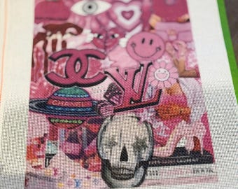 May include: A needlepoint canvas featuring a collage of pink-toned imagery, including a skull, a smiley face, a planet, and luxury brand logos. The design incorporates various graphic elements like hearts and an eye, all printed on a white mesh grid.