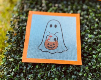 May include: A square needlepoint canvas featuring a white ghost outline on a light blue background. The ghost is holding an orange jack-o'-lantern bucket containing a small white chicken with a red comb. The canvas has a solid orange border.