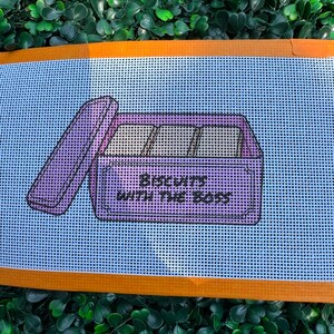 May include: A needlepoint canvas featuring a light blue background with an orange border. The design depicts a purple rectangular tin with an open lid, containing three square biscuits. The text "BISCUITS WITH THE BOSS" is printed in black.