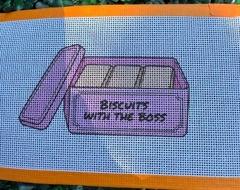 May include: A needlepoint canvas featuring a light blue background with an orange border. The design depicts a purple rectangular tin with an open lid, containing three square biscuits. The text "BISCUITS WITH THE BOSS" is printed in black.