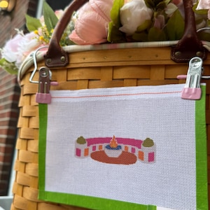 May include: A rectangular white needlepoint canvas featuring a small, colorful embroidered design of a pink and orange structure with a central flame. The canvas has bright green fabric borders and a small white label reading Sunset Stitching.