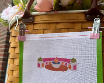 May include: A rectangular white needlepoint canvas featuring a small, colorful embroidered design of a pink and orange structure with a central flame. The canvas has bright green fabric borders and a small white label reading Sunset Stitching.
