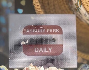 May include: A square white needlepoint canvas featuring a maroon rectangular design with rounded corners. The text "ASBURY PARK" is printed in white at the top, followed by a black horizontal wavy line, and the word "DAILY" in white at the bottom.