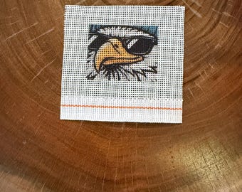 May include: A square piece of white needlepoint canvas featuring a printed graphic of a bald eagle wearing black sunglasses. The eagle has a yellow beak and white feathers. A single horizontal orange thread line runs across the bottom.