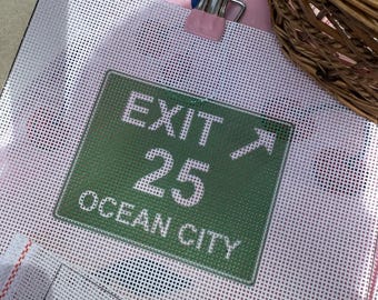 May include: A white needlepoint canvas featuring a green rectangular sign design with white text that reads EXIT 25 OCEAN CITY and an arrow pointing diagonally upward. A silver metal clip with a pink accent is attached to the top of the canvas.
