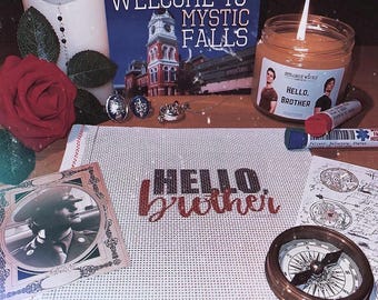 May include: A cross-stitch fabric piece with the text HELLO brother in black and red thread, surrounded by a candle labeled HELLO, BROTHER, a Welcome to Mystic Falls sign, a red rose, two rings, a locket, a compass, and a photo of a person in uniform.