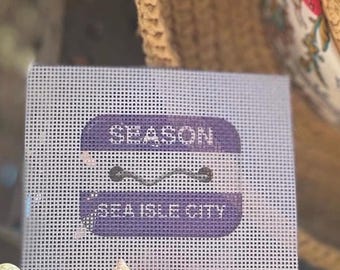 May include: A rectangular white needlepoint canvas featuring a purple graphic design with rounded corners. The design includes the text SEASON at the top, a horizontal wavy line in the center, and the text SEA ISLE CITY at the bottom.