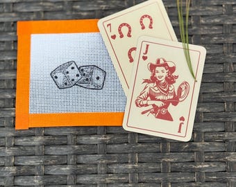 May include: A square needlepoint canvas with an orange border featuring a black printed graphic of two dice, accompanied by two playing cards. One card shows a red horseshoe and the number seven, while the other depicts a cowgirl with a lasso.