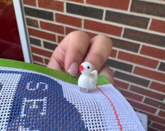 May include: A small white duck-shaped needle minder with a bright red beak and a black eye, attached to a piece of white needlepoint canvas featuring a blue grid pattern and the partial text "SEA" stitched in white thread on the fabric.