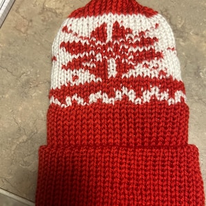 May include: Red knit beanie with a white knit pattern on the top. The pattern is a geometric design.