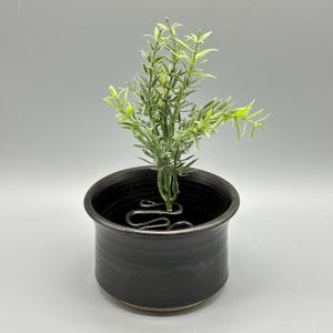 Może przedstawiać: Mała, czarna ceramiczna donica o teksturowanej powierzchni, w której znajduje się zielono-żółta sztuczna roślina. Roślina jest zabezpieczona ozdobnym metalowym wspornikiem wewnątrz donicy. Donica ma okrągły kształt i jasną podstawę.