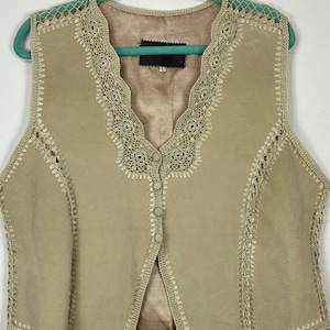 May include: A beige suede vest featuring intricate crochet trim along the V-neckline, armholes, and hem. The garment includes a front button closure with four matching fabric-covered buttons and a smooth, tonal interior lining.