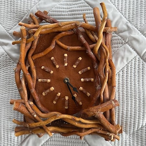 Könnte beinhalten: Eine quadratische Wanduhr mit braunem Lederzifferblatt und einem Rahmen aus verschlungenen Zweigen. Das Zifferblatt hat kleine Holzmarkierungen für die Stunden und schwarze Zeiger. Das Design hat eine rustikale, natürliche Ästhetik.