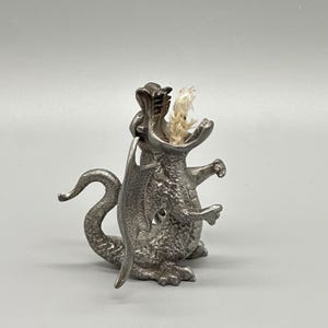 May include: A silver-colored dragon-shaped oil lamp with a wide open mouth and a wick. The dragon has wings and a long tail. The lamp is made of metal and sits on a white surface.