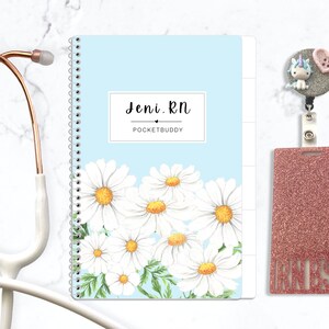 May include: A light blue spiral-bound notebook with a white daisy floral pattern, featuring the text Jeni. RN POCKETBUDDY, a rose gold stethoscope, and a glittery rose gold badge reel with a unicorn and heart charm and the text RN BSN.