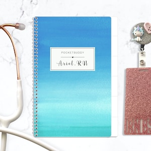 May include: A spiral-bound notebook with a blue watercolor gradient cover featuring the text POCKETBUDDY and Ariel, RN, a rose gold stethoscope, and a glittery pink badge reel with a unicorn and heart charm and a matching glittery ID holder.