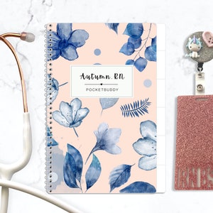 May include: A spiral-bound notebook with a light peach cover featuring blue floral patterns and the text Autumn.RN POCKETBUDDY, a rose gold stethoscope, and a glittery pink badge holder with a unicorn and heart charm and the text RNBSN.
