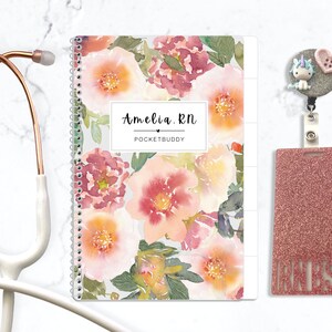 May include: A rose gold stethoscope, a spiral-bound notebook with a watercolor floral pattern featuring the text Amelia. RN POCKETBUDDY, and a glittery rose gold badge reel with a unicorn and heart button charm and a matching glittery ID holder.