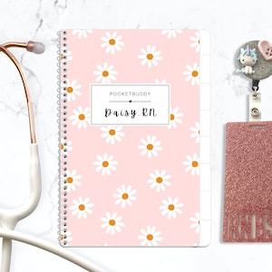 May include: A rose gold stethoscope, a spiral-bound notebook with a pink daisy pattern featuring the text POCKETBUDDY Daisy.RN, and a glittery rose gold badge reel with a unicorn and heart charm attached to a matching glittery ID holder.